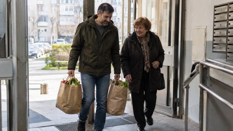 Un bărbat ajută o femeie în vârstă, cărând două sacoșe cu cumpărături la intrarea în bloc.