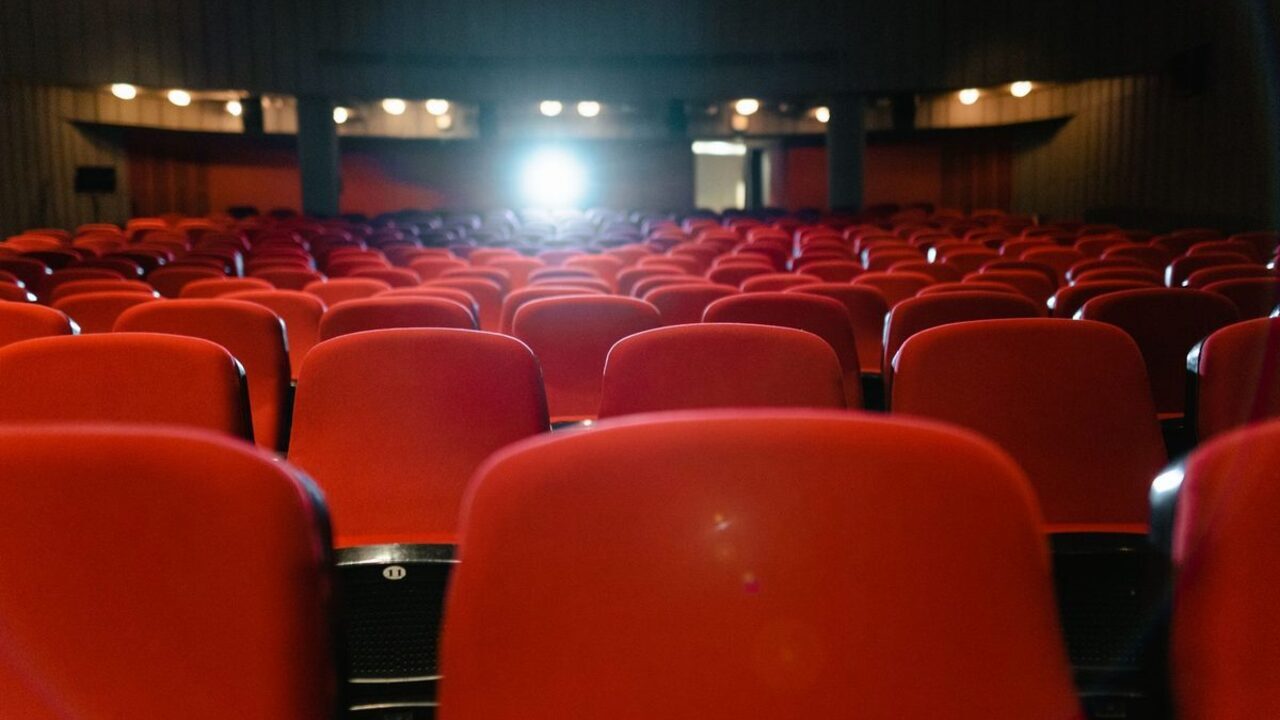 Rânduri de scaune roșii într-o sală de cinema iluminată discret de lumina proiectorului.