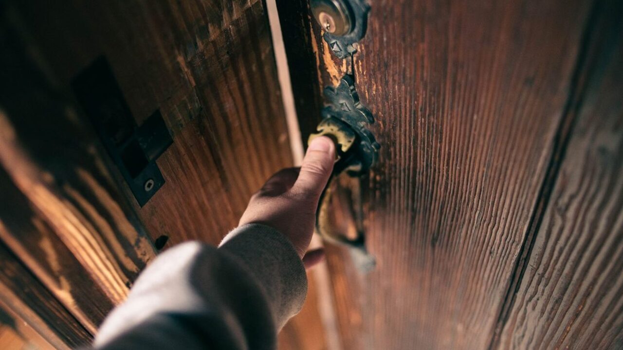 O mână acționează clanța metalică pentru a deschide o ușă masivă din lemn.