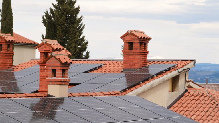 Panouri fotovoltaice montate pe acoperișuri din țiglă roșie, cu coșuri de cărămidă, sub un cer înnorat.