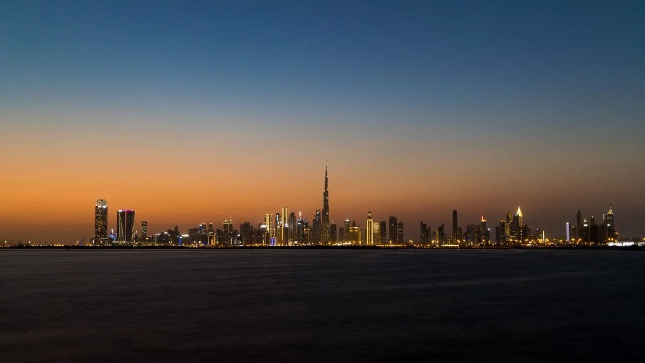 Linia orizontului din Dubai cu zgârie-nori luminați și turnul Burj Khalifa la apus peste apă.