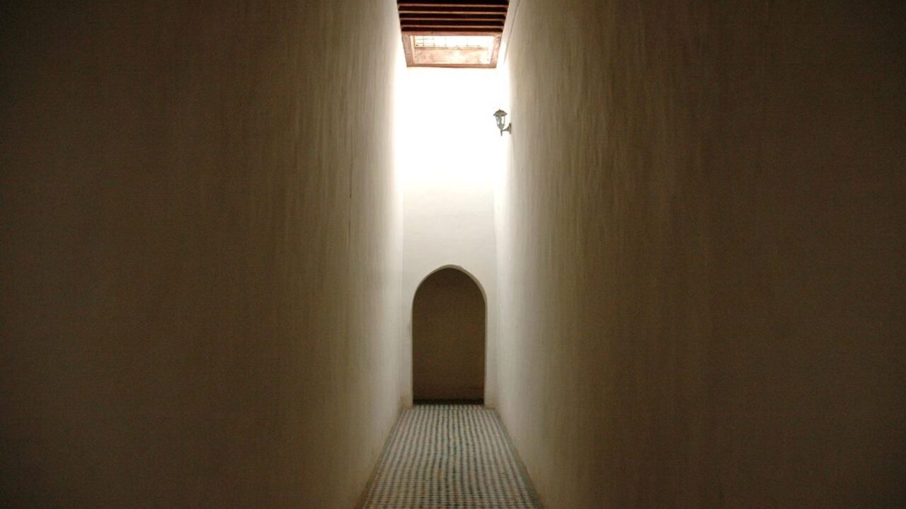 Un pasaj interior îngust delimitat de pereți înalți duce spre o arcadă luminoasă.