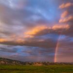 Curcubeu spectaculos printre nori dramatici la apus, într-un peisaj de țară luxuriant