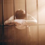 Bărbat privind printre gratiile unei închisori cu raze de soare, simbolizând speranța și dorința de libertate în vis