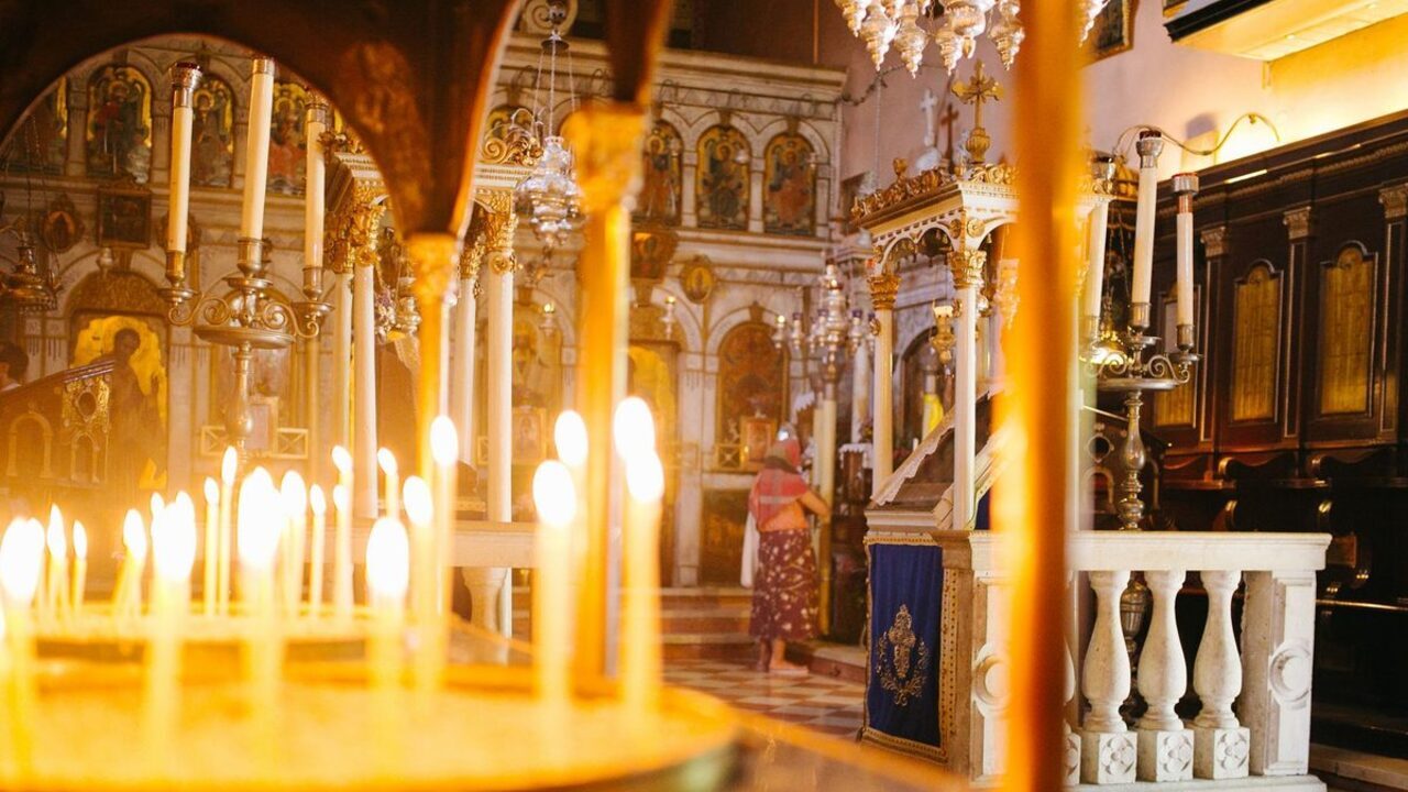 Interiorul unei biserici ortodoxe cu lumânări aprinse în prim-plan și catapeteasma în fundal.