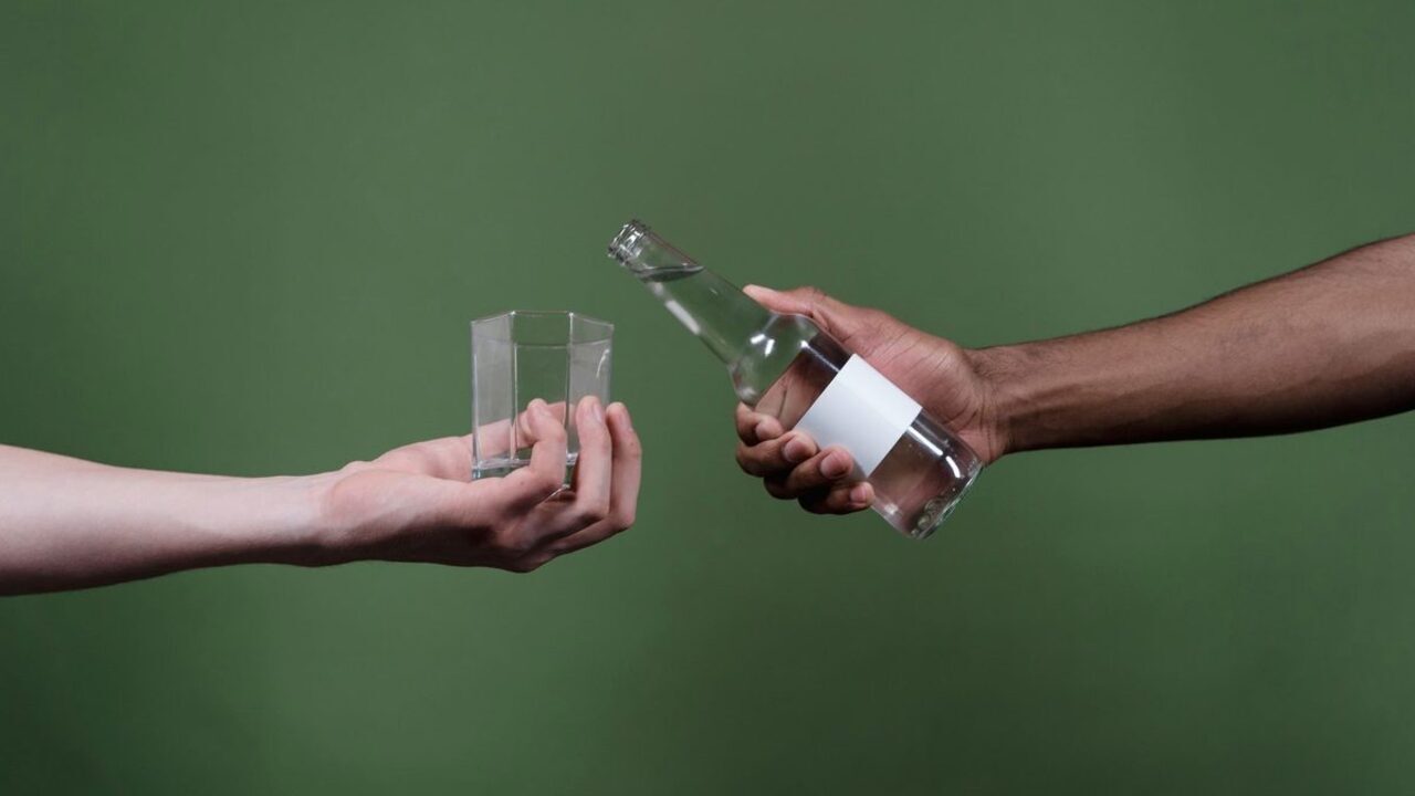 O mână toarnă apă dintr-o sticlă transparentă într-un pahar ținut de o altă persoană.