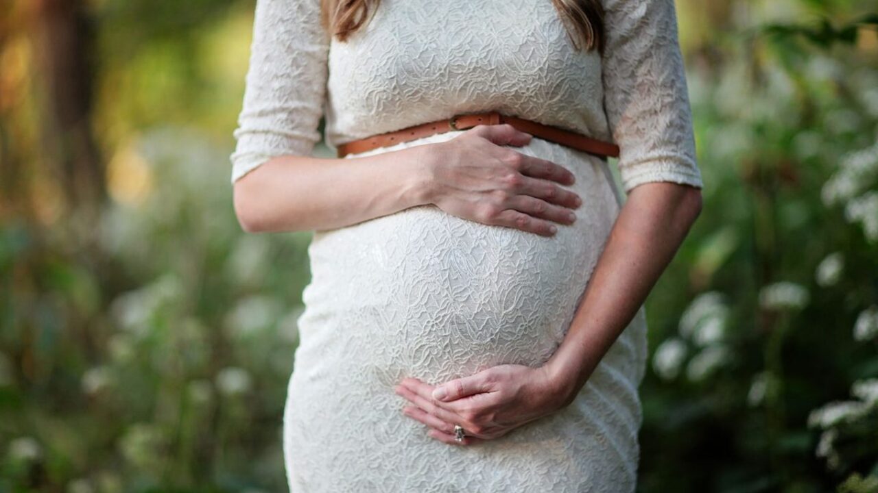 O femeie însărcinată într-o rochie albă își sprijină mâna pe burtă într-un cadru natural.