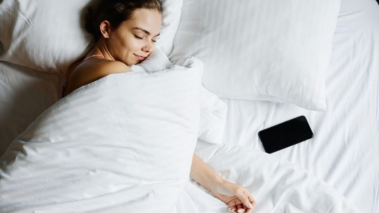 O persoană stă întinsă pe un pat cu așternuturi albe lângă un telefon mobil.