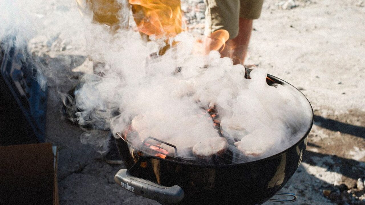 Nori de fum alb și dens se ridică deasupra bucăților de carne așezate pe un grătar cu cărbuni.