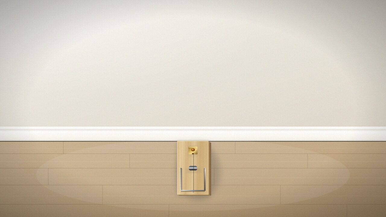 O capcană clasică din lemn pentru șoareci, așezată perpendicular pe plinta albă a unui perete, pe o podea din parchet, în lumină naturală de interior.