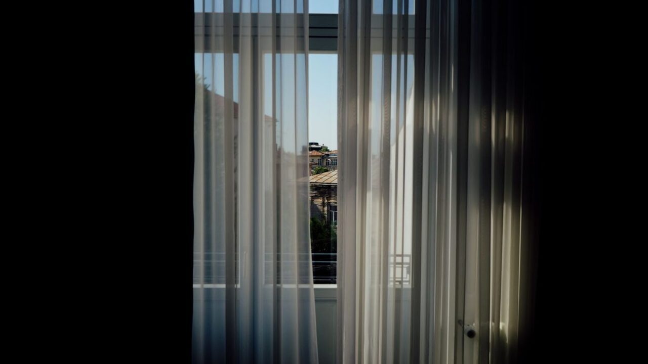 Draperii albe vaporoase filtrează lumina naturală la o fereastră deschisă spre oraș.