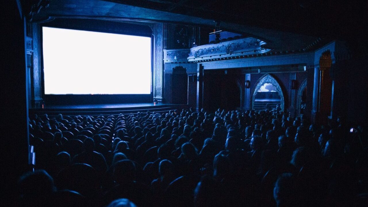 O sală mare de cinema plină de spectatori privește spre un ecran uriaș luminos.