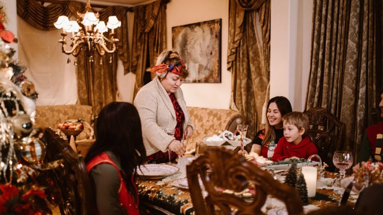 Membrii unei familii extinse stau adunați în jurul mesei în timp ce o femeie servește mâncarea.