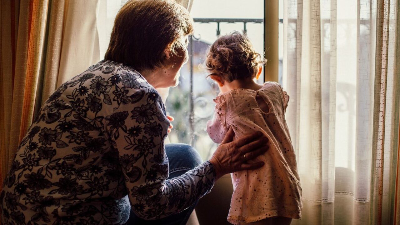O bunică și o fetiță privesc împreună pe fereastră din interiorul unei camere luminoase.