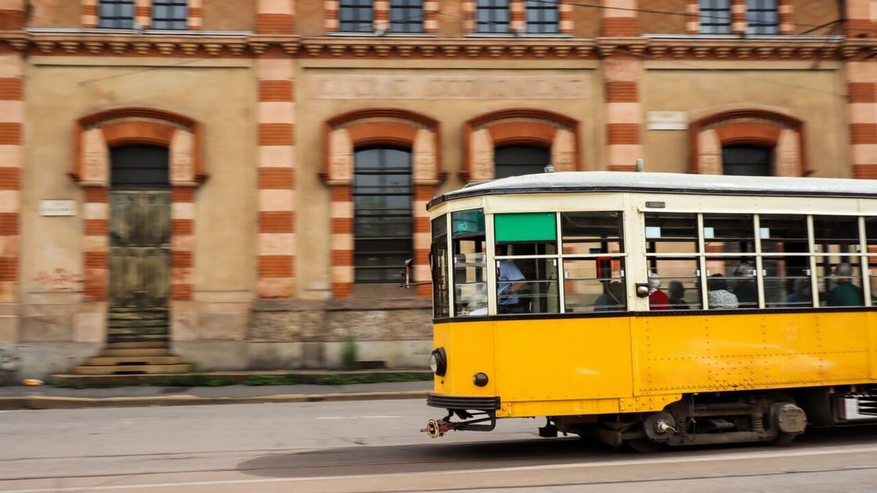 Un tramvai galben de epocă rulează cu viteză pe șine pe lângă o clădire istorică.